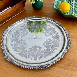 May include: A silver serving tray with an ornate, embossed design. The tray has a raised rim and a centre inscription that reads "To Alfred Bell, Esq. who won the 1924-1925 Championship Cricket Match".
