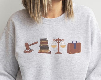 Puede incluir: Una sudadera gris con un diseño gráfico de un martillo, una pila de libros, una balanza de la justicia y un maletín. El diseño es en marrón, azul y amarillo.