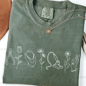 May include: Moss green t-shirt with white line art of tools, flowers, and jewelry. A gold necklace with a pendant rests on the shirt. The text "MOSS" is at the bottom left. The shirt is a Comfort Colors brand.