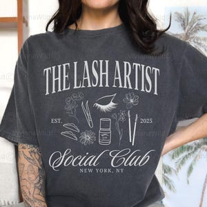 May include: Pepper-colored t-shirt with the text "THE LASH ARTIST Social Club NEW YORK, NY" in white. The design includes lash tools, flowers, and the year 2025. A casual, comfortable tee for lash artists.