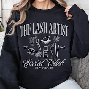 Peut inclure: Sweat-shirt noir avec l'inscription blanche "THE LASH ARTIST Social Club NEW YORK, NY". Le motif comprend des outils pour les cils, des fleurs et l'année 2025. Un vêtement confortable pour les artistes des cils.