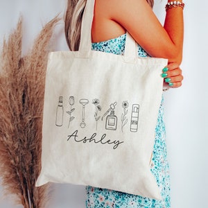Peut inclure: Un sac fourre-tout en toile blanche avec des dessins au trait noir de produits de beaut&eacute; et de fleurs. Le nom "Ashley" est imprim&eacute; en cursive sous les dessins.