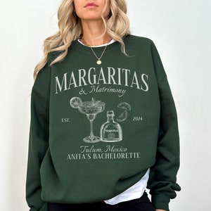 May include: Forest green sweatshirt with white text that reads "Margaritas & Matrimony" with a margarita glass and tequila bottle illustration. The text also includes "EST. 2024" and "Tulum, Mexico Anita's Bachelorette".