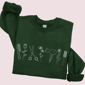 Puede incluir: Una sudadera verde oscuro con un contorno blanco de un peine, unas tijeras, un secador de pelo y un cepillo para el pelo con flores entre cada elemento.