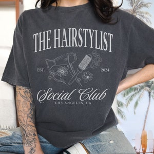 May include: Pepper-colored t-shirt with a white graphic design. The design includes the text "THE HAIRSTYLIST Social Club LOS ANGELES, CA" along with illustrations of a hair dryer, brush, and flowers. The year "2024" is also included.