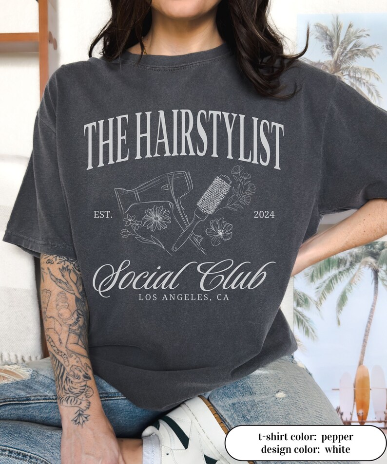 May include: Pepper-colored t-shirt with a white graphic design. The design includes the text "THE HAIRSTYLIST Social Club LOS ANGELES, CA" along with illustrations of a hair dryer, brush, and flowers. The year "2024" is also included.