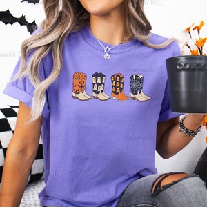 Puede incluir: Una camiseta violeta con un gr&aacute;fico de cuatro botas de vaquero con tem&aacute;tica de Halloween. Las botas est&aacute;n decoradas con calabazas, fantasmas, caramelos y telara&ntilde;as. El texto "Violet" es visible en la esquina superior derecha.