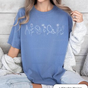May include: Blue jean colored t-shirt with a white line drawing of tools and flowers. The text "t-shirt color: blue jean" is at the bottom of the image. The shirt is a comfortable, casual style.