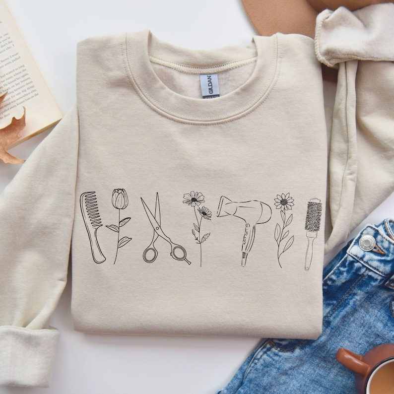 Puede incluir: Una sudadera beige con un dise&ntilde;o gr&aacute;fico de herramientas de peluquer&iacute;a, que incluyen un peine, unas tijeras, un secador de pelo y un cepillo, todos con detalles florales.