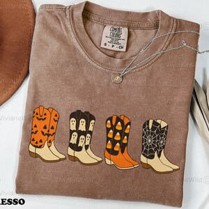 Puede incluir: Camiseta Comfort Colors color espresso con cuatro dise&ntilde;os de botas de vaquero con tem&aacute;tica de Halloween. Las botas son naranjas con caras de calabaza, negras con fantasmas, naranjas con caramelos y negras con una telara&ntilde;a. Tambi&eacute;n se ve un collar plateado.