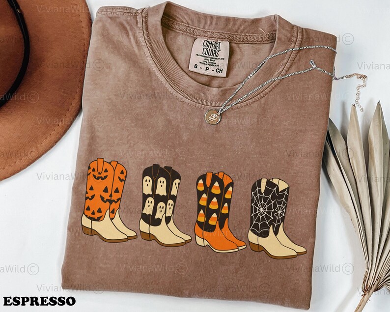 Puede incluir: Camiseta Comfort Colors color espresso con cuatro dise&ntilde;os de botas de vaquero con tem&aacute;tica de Halloween. Las botas son naranjas con caras de calabaza, negras con fantasmas, naranjas con caramelos y negras con una telara&ntilde;a. Tambi&eacute;n se ve un collar plateado.
