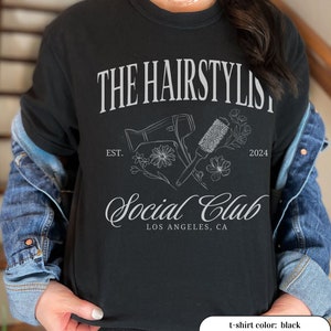 May include: Black t-shirt with the text "THE HAIRSTYLIST Social Club LOS ANGELES, CA" in white. The design includes a hair dryer, round brush, flowers, and the year 2024. The shirt is a crew neck style.