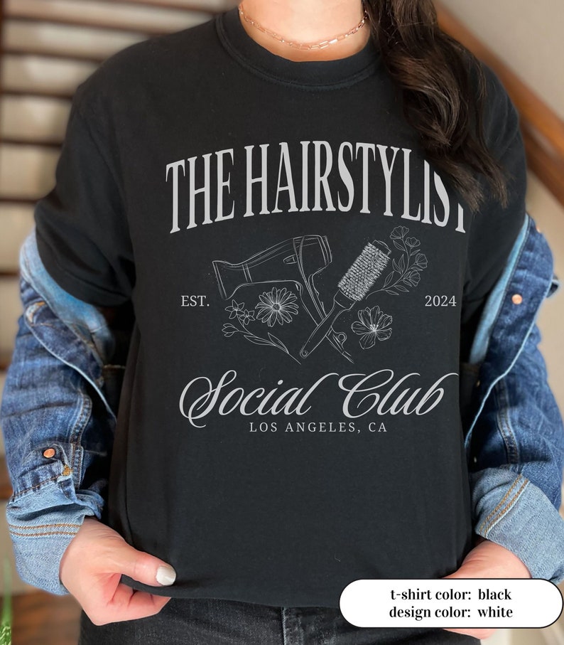 May include: Black t-shirt with the text "THE HAIRSTYLIST Social Club LOS ANGELES, CA" in white. The design includes a hair dryer, round brush, flowers, and the year 2024. The shirt is a crew neck style.