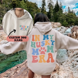 May include: A beige hoodie with the text "In My Husky Mom Era" in rainbow colors on the back. The front of the hoodie has a smaller version of the text with a paw print and a space for a custom dog name.