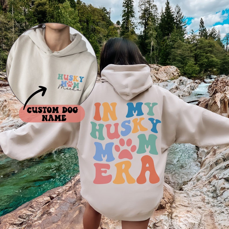 May include: A beige hoodie with the text "In My Husky Mom Era" in rainbow colors on the back. The front of the hoodie has a smaller version of the text with a paw print and a space for a custom dog name.