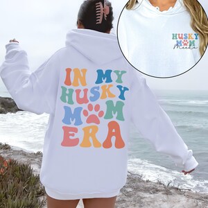 May include: White hoodie with a rainbow colored graphic that says "In My Husky Mom Era" on the back. The front of the hoodie has a small graphic that says "Husky Mom" with a paw print and the name "Meika" written in cursive.