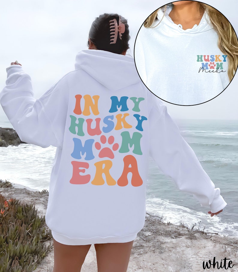 May include: White hoodie with a rainbow colored graphic that says "In My Husky Mom Era" on the back. The front of the hoodie has a small graphic that says "Husky Mom" with a paw print and the name "Meika" written in cursive.