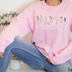 Puede incluir: Una sudadera rosa claro con un dise&ntilde;o gr&aacute;fico de herramientas de peluquer&iacute;a, incluyendo tijeras, un peine, un secador de pelo y un cepillo. Las herramientas est&aacute;n delineadas en negro y est&aacute;n dispuestas en una fila a trav&eacute;s de la parte delantera de la sudadera.