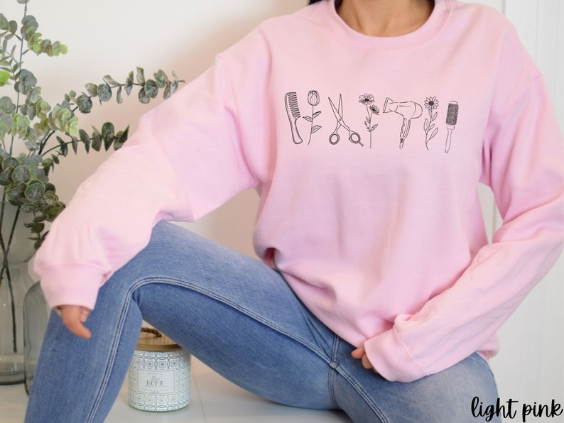 Puede incluir: Una sudadera rosa claro con un dise&ntilde;o gr&aacute;fico de herramientas de peluquer&iacute;a, incluyendo tijeras, un peine, un secador de pelo y un cepillo. Las herramientas est&aacute;n delineadas en negro y est&aacute;n dispuestas en una fila a trav&eacute;s de la parte delantera de la sudadera.