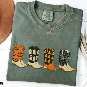 Puede incluir: Camiseta Comfort Colors verde musgo con una fila de botas de vaquero con tem&aacute;tica de Halloween. Las botas son naranjas con caras de calabaza, negras con fantasmas, naranjas con caramelos y negras con telara&ntilde;as. Tambi&eacute;n se ve un collar dorado y un sombrero marr&oacute;n.