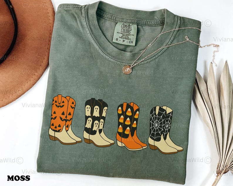 Puede incluir: Camiseta Comfort Colors verde musgo con una fila de botas de vaquero con tem&aacute;tica de Halloween. Las botas son naranjas con caras de calabaza, negras con fantasmas, naranjas con caramelos y negras con telara&ntilde;as. Tambi&eacute;n se ve un collar dorado y un sombrero marr&oacute;n.