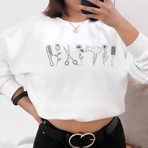 Puede incluir: Sudadera blanca con un contorno negro de herramientas de peluquer&iacute;a, que incluyen un peine, unas tijeras, un secador de pelo y un cepillo redondo. Las herramientas est&aacute;n dispuestas horizontalmente en la parte delantera de la sudadera.