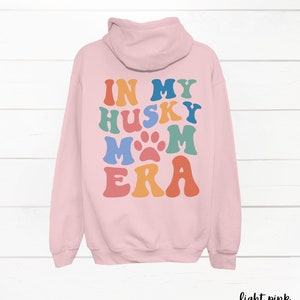 May include: Light pink hooded sweatshirt with the text "In My Husky Mom Era" in rainbow colors with a paw print in the middle of the word "Mom".