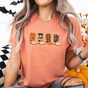 Puede incluir: Camiseta color terracota con una fila de botas de vaquero de dibujos animados con dise&ntilde;os de Halloween. Las botas incluyen calabazas, fantasmas, caramelos de ma&iacute;z y telara&ntilde;as. El texto "Terracota" est&aacute; en la esquina superior derecha.