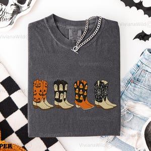 Puede incluir: Camiseta gris oscuro Comfort Colors con cuatro dise&ntilde;os de botas de vaquero con tem&aacute;tica de Halloween. Las botas son naranjas, negras y blancas, con calabazas, fantasmas, caramelos y telara&ntilde;as. Un collar de cadena plateada est&aacute; sobre la camisa.