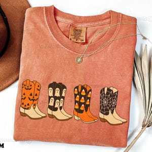 Puede incluir: Una camiseta de color coral con cuatro dise&ntilde;os de botas de vaquero con tem&aacute;tica de Halloween. Las botas est&aacute;n decoradas con calabazas, fantasmas, caramelos de ma&iacute;z y telara&ntilde;as. La camiseta tiene cuello redondo y una etiqueta peque&ntilde;a que dice "Comfort Colors".
