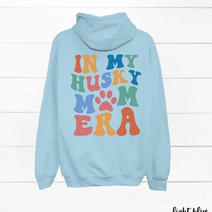 May include: Light blue hooded sweatshirt with the text "In My Husky Mom Era" in rainbow colors with a paw print in the middle of the word "Mom".
