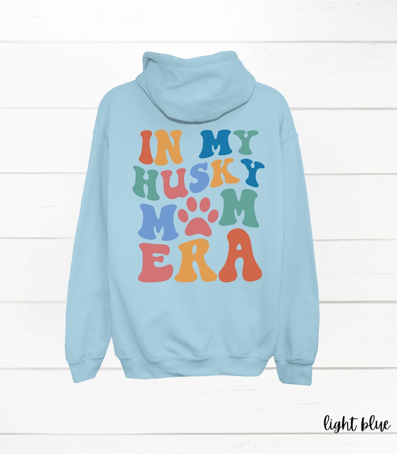 May include: Light blue hooded sweatshirt with the text "In My Husky Mom Era" in rainbow colors with a paw print in the middle of the word "Mom".