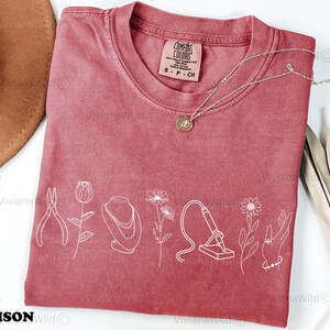 May include: A dusty rose Comfort Colors t-shirt with white line art of jewelry tools and flowers. A gold necklace with a pendant rests on the shirt. The shirt has a tag that reads "S P CH". The brand name "Crimson Wild" is at the bottom.