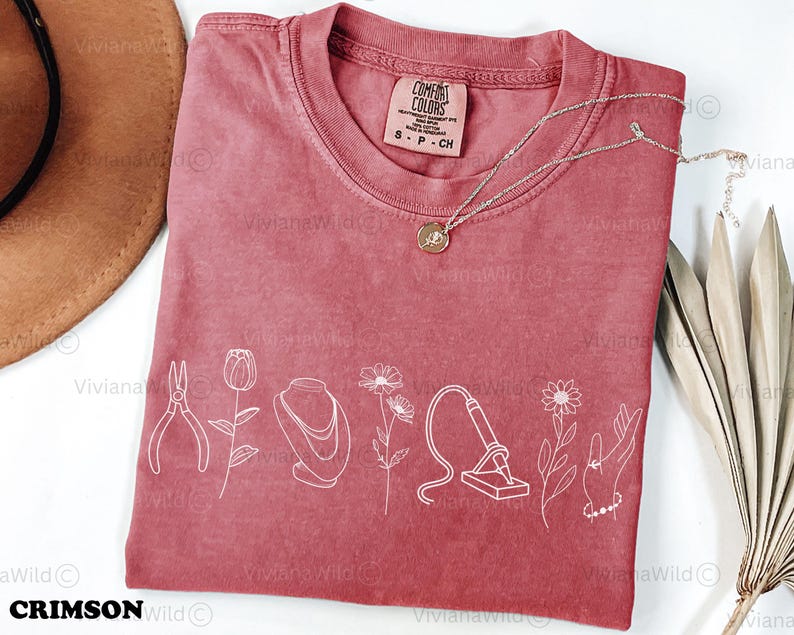 May include: A dusty rose Comfort Colors t-shirt with white line art of jewelry tools and flowers. A gold necklace with a pendant rests on the shirt. The shirt has a tag that reads "S P CH". The brand name "Crimson Wild" is at the bottom.