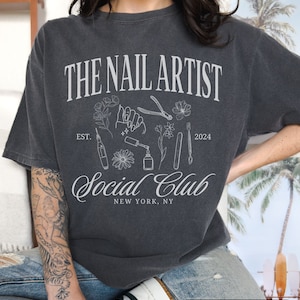 May include: A dark grey t-shirt with a white design that says "The Nail Artist Social Club" with a stylised image of nail art tools and flowers. The text "Est. 2024" and "New York, NY" are also included in the design.