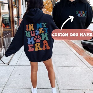 May include: Black hooded sweatshirt with the text "In My Husky Mom Era" in rainbow colors on the back. The front of the sweatshirt has the text "Husky Mom" with a paw print and space for a custom dog name.