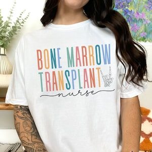May include: White t-shirt with the text "BONE MARROW TRANSPLANT nurse" in rainbow colors. A skeleton hand is making the "I love you" sign language.