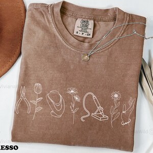 May include: Espresso-colored Comfort Colors t-shirt with a white line drawing of jewelry-making tools and flowers. A silver necklace with a pendant is on the shirt. The shirt has the text "ESPRESSO" at the bottom.