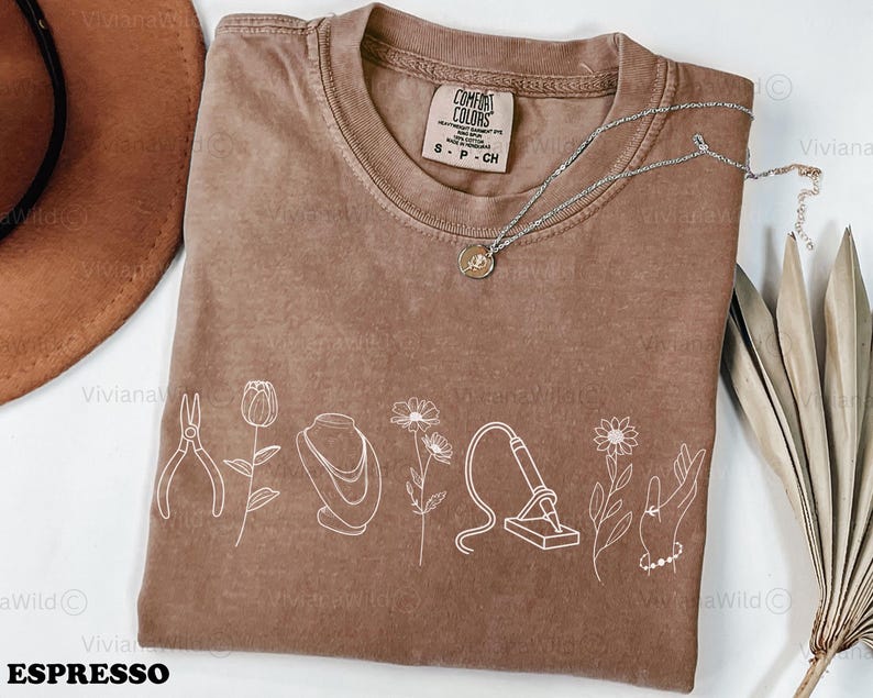 May include: Espresso-colored Comfort Colors t-shirt with a white line drawing of jewelry-making tools and flowers. A silver necklace with a pendant is on the shirt. The shirt has the text "ESPRESSO" at the bottom.