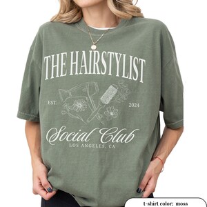 May include: Moss green t-shirt with the text "THE HAIRSTYLIST Social Club" in white. The design includes a hair dryer, brush, and floral elements. The text "EST. 2024" and "LOS ANGELES, CA" are also included in the design.