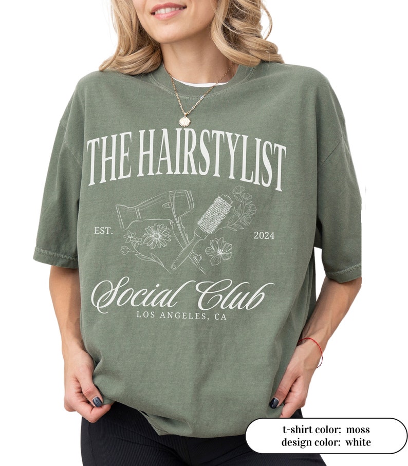 May include: Moss green t-shirt with the text "THE HAIRSTYLIST Social Club" in white. The design includes a hair dryer, brush, and floral elements. The text "EST. 2024" and "LOS ANGELES, CA" are also included in the design.