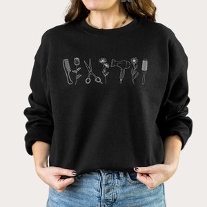 Puede incluir: Sudadera negra con un contorno blanco de herramientas de peluquer&iacute;a y flores. El texto "Black" es visible en la esquina inferior derecha de la imagen.