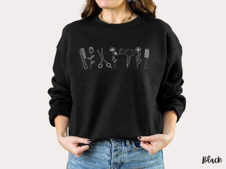 Puede incluir: Sudadera negra con un contorno blanco de herramientas de peluquer&iacute;a y flores. El texto "Black" es visible en la esquina inferior derecha de la imagen.