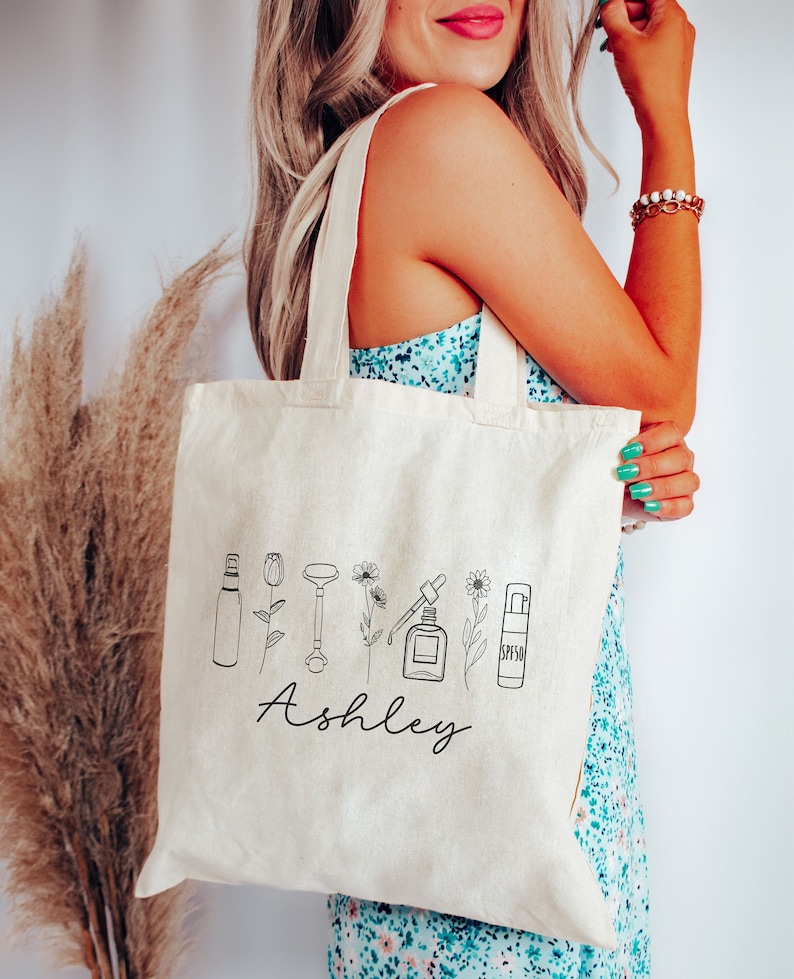 Peut inclure: Un sac fourre-tout en toile blanche avec des dessins au trait noir de produits de beaut&eacute; et de fleurs. Le nom "Ashley" est imprim&eacute; en cursive sous les dessins.
