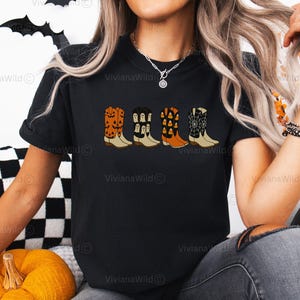 Puede incluir: Camiseta negra con un dise&ntilde;o de cuatro botas de vaquero decoradas con motivos de Halloween. Las botas presentan calabazas, fantasmas, caramelos de ma&iacute;z y telara&ntilde;as. La camiseta tiene cuello redondo y la palabra "Black" est&aacute; en la parte inferior.