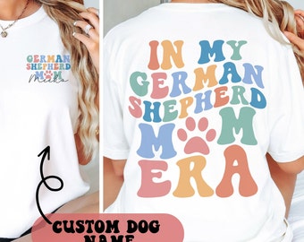 May include: White t-shirt with a colorful graphic that says "In My German Shepherd Mom Era" with a paw print in the middle. The shirt has a smaller graphic on the front that says "German Shepherd Mom" with a paw print and the name "Meeko".