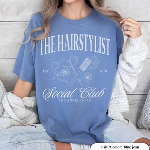 May include: Blue jean t-shirt with white text and graphics. The design includes the words "THE HAIRSTYLIST SOCIAL CLUB" with "EST. 2024" and "LOS ANGELES, CA" below. The graphic features a hair dryer, brush, and flowers.