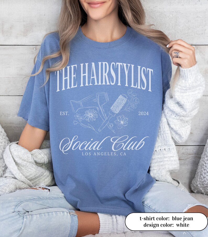 May include: Blue jean t-shirt with white text and graphics. The design includes the words "THE HAIRSTYLIST SOCIAL CLUB" with "EST. 2024" and "LOS ANGELES, CA" below. The graphic features a hair dryer, brush, and flowers.