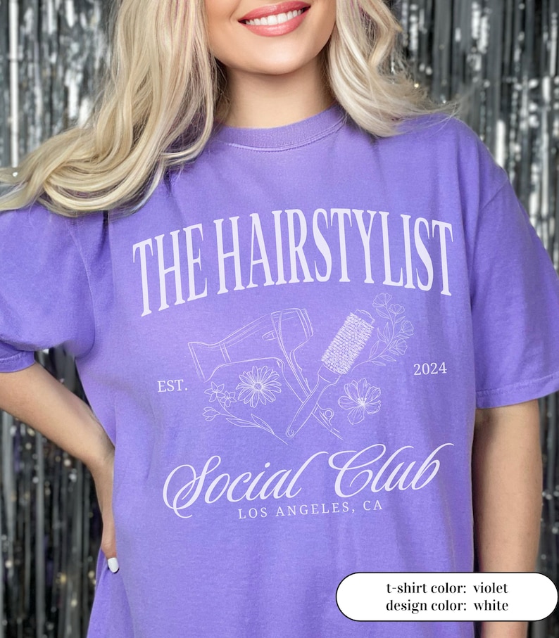 May include: A violet t-shirt with the text "THE HAIRSTYLIST Social Club LOS ANGELES, CA" in white. The design includes a hair dryer, brush, flowers, and the year 2024. The shirt is a solid color.