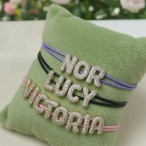 Op de afbeelding: Drie gepersonaliseerde armbanden met de namen "NOR", "LUCY" en "VICTORIA" in sprankelende, met edelstenen bezette letters. Elke naam is geregen op een verschillend gekleurd koord: paars, zwart en roze. De armbanden liggen op een groen kussen.